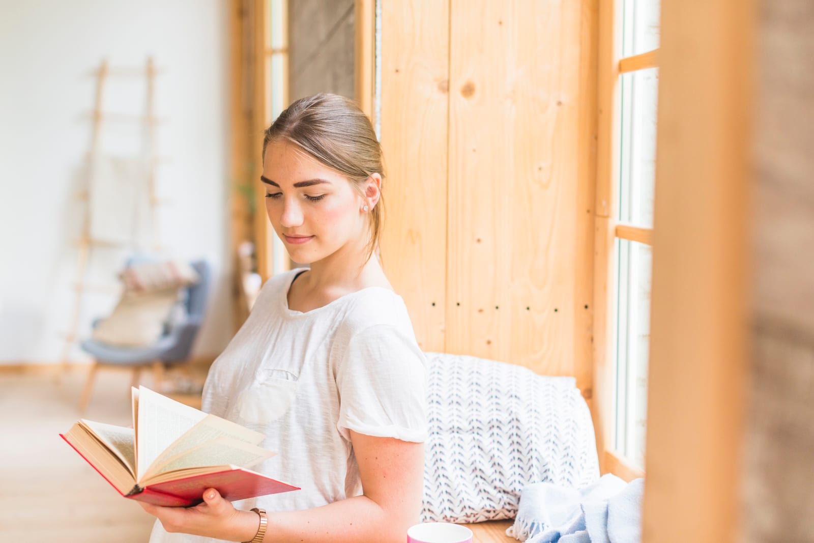 Leitora em vestido claro, de pé junto a uma janela, absorta em um livro de capa vermelha