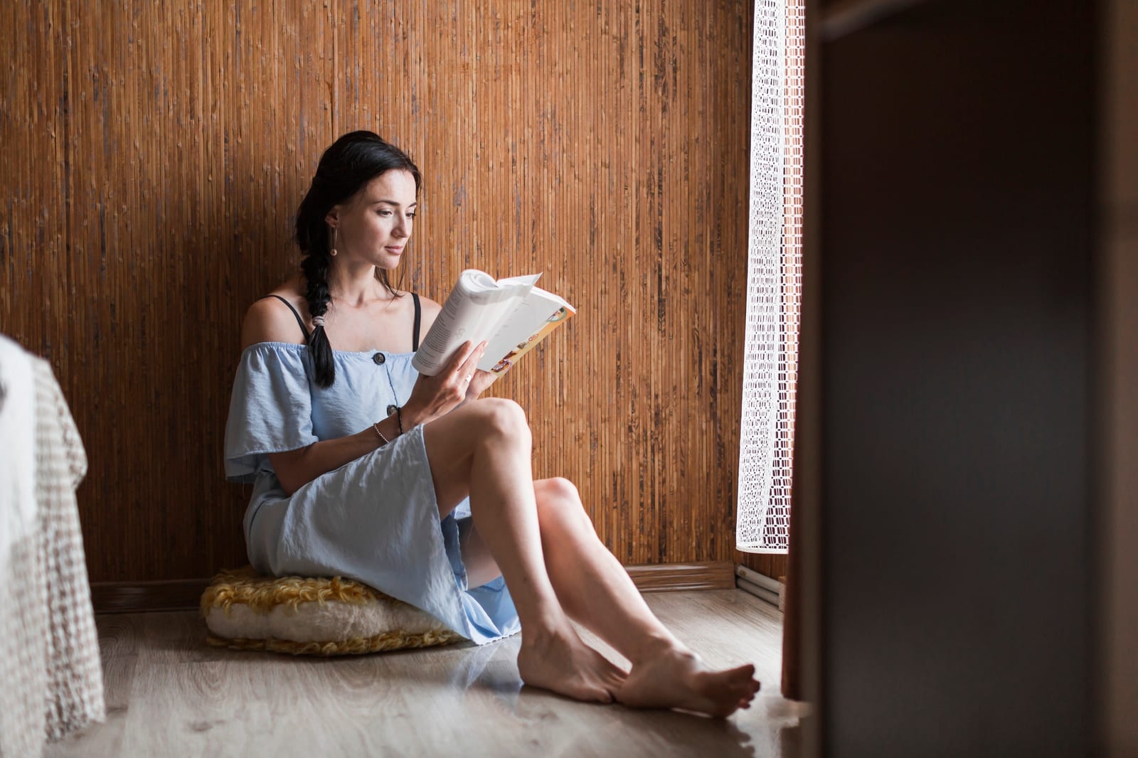 Leitora sentada em uma almofada no chão, perto de uma janela, concentrada em um livro aberto