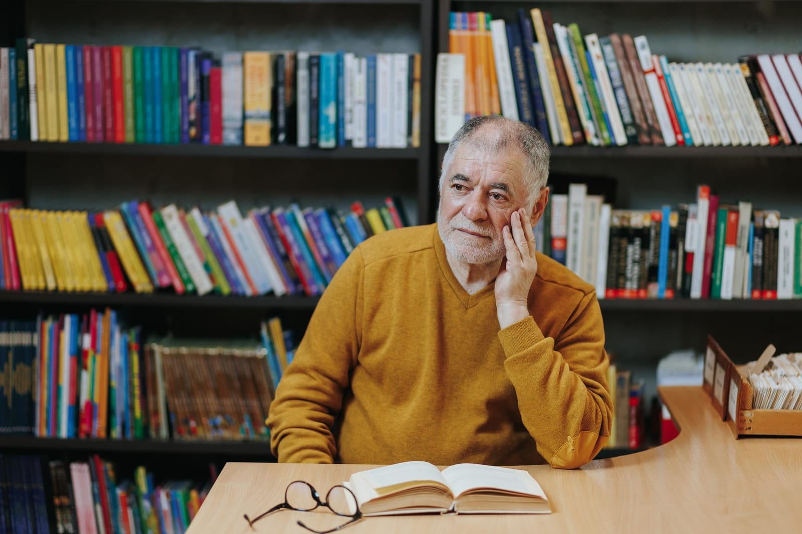 Leitor sentado em uma biblioteca com um livro aberto sobre a mesa e óculos ao lado